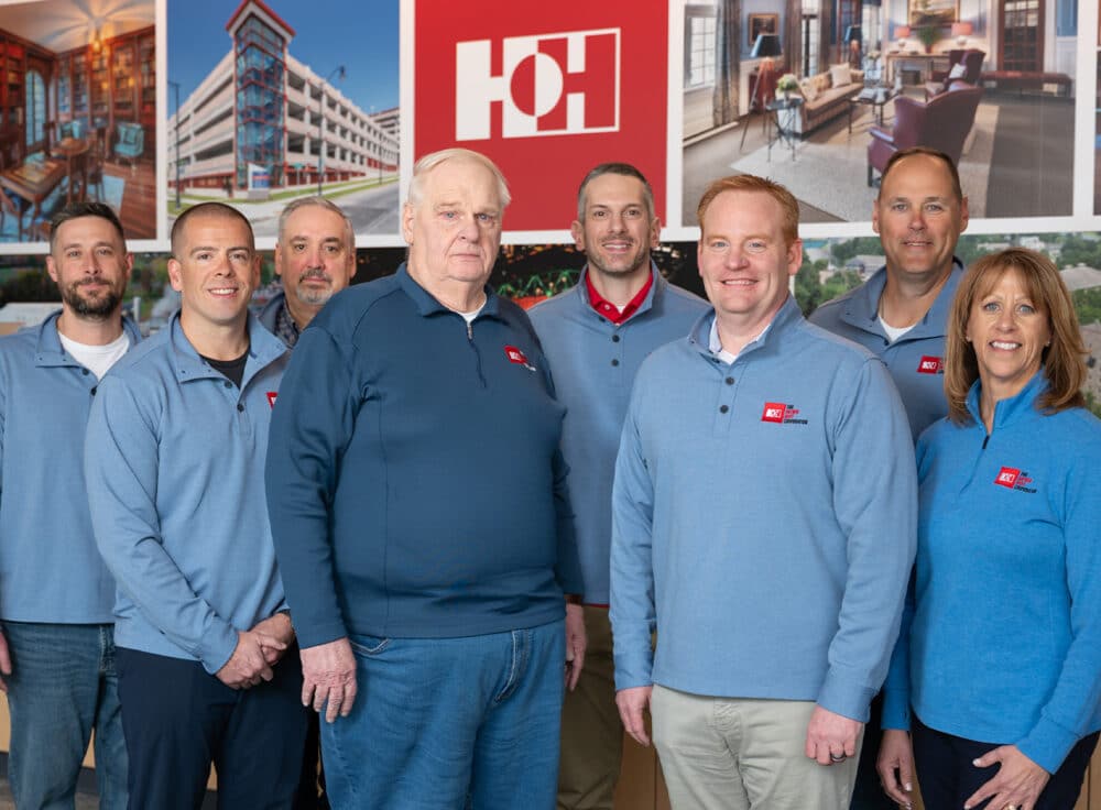 Members of Hayner Hoyt's senior leadership team wearing blue sweaters standing proudly in front of a wall showing recent construction and build projects.