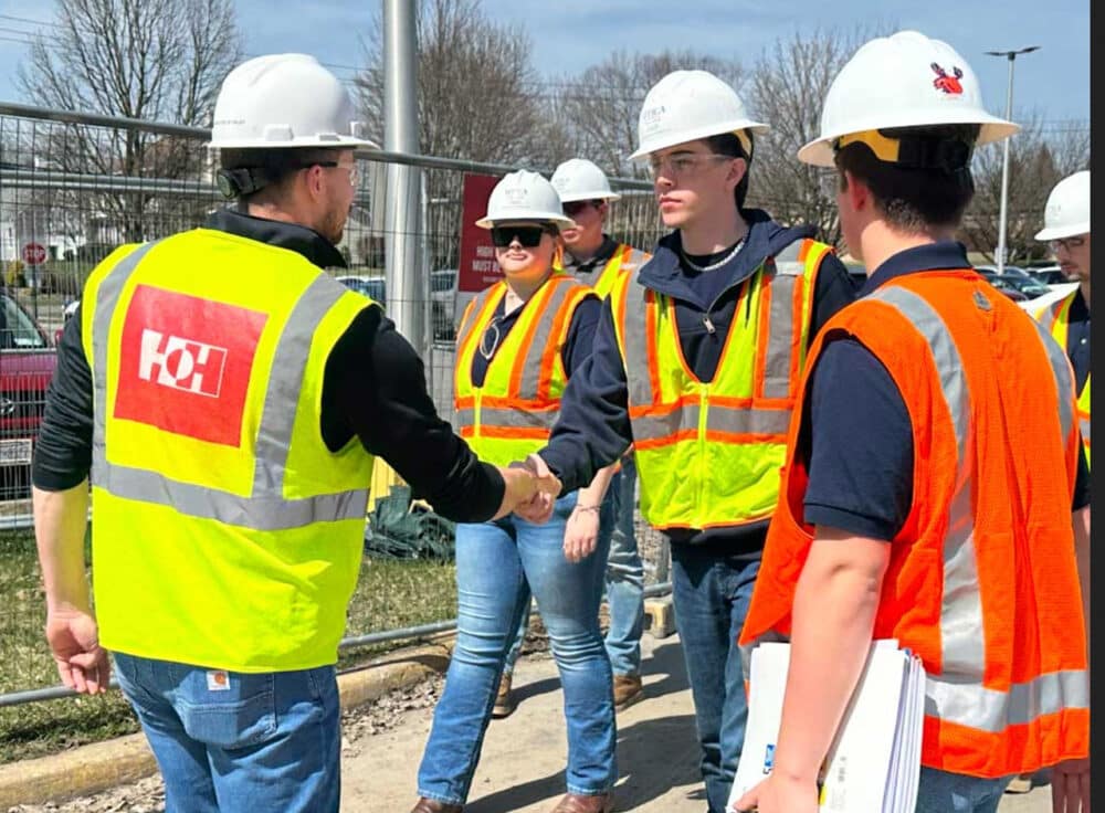 Team members wearing white hard hats and yellow high visibility vests shaking hands on a job site.